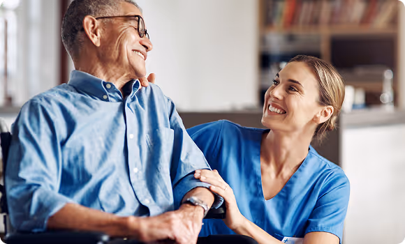 Smiling female caregiver in blue scrubs holding the arm of a cheerful elderly man in a wheelchair.