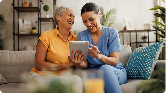 A healthcare worker in blue scrubs showing a tablet to a smiling elderly woman on a couch.