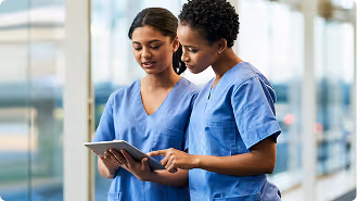 Two female healthcare professionals in blue scrubs looking at and discussing a tablet device.