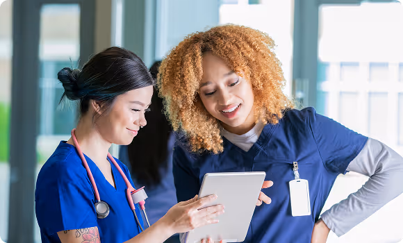 Two smiling female healthcare professionals in scrubs looking at a tablet together.