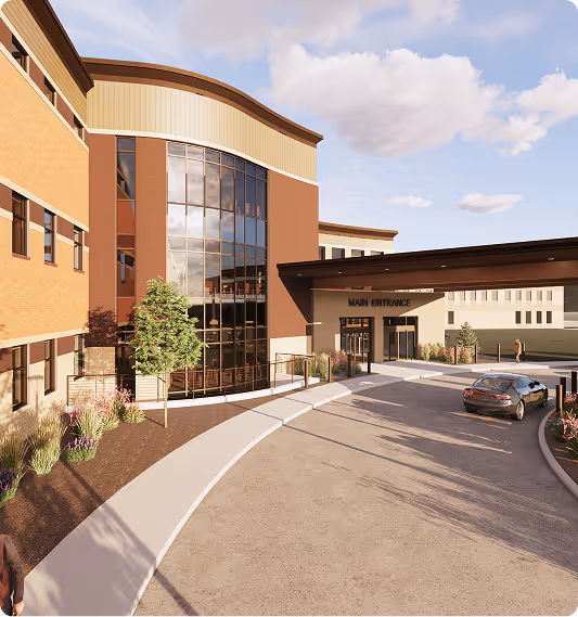 Modern building with large curved glass windows, a main entrance canopy, landscaped surroundings, and a car parked on the driveway.