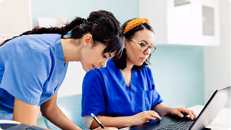 Two healthcare professionals in blue scrubs working together, one writing notes and the other using a laptop in a clinical setting.