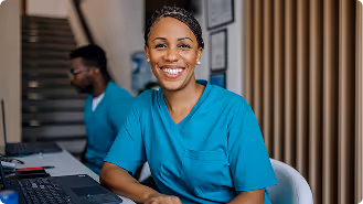 Smiling female healthcare worker in teal scrubs sitting at a desk with a computer.