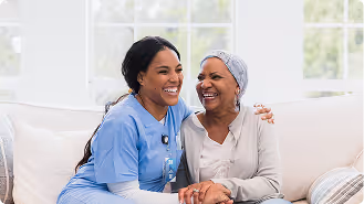 Smiling nurse in blue scrubs sitting on a couch with an elderly woman wearing a headscarf, holding hands and sharing a joyful moment.