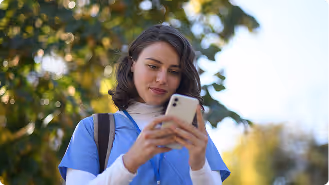 Young woman in blue scrubs and a backpack looking at her smartphone outdoors with trees in the background.