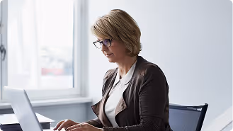 Woman with short blonde hair and glasses typing on a laptop at a desk by a window.
