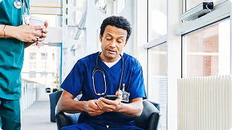 Male healthcare professional in blue scrubs sitting and looking at his phone, with another person standing nearby in a hospital hallway.