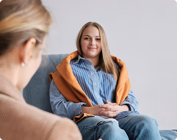 Young woman in a striped shirt and orange sweater sitting on a chair, smiling during a conversation.
