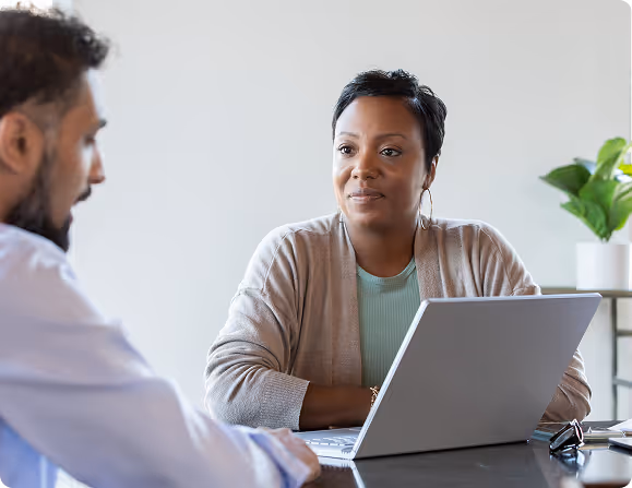 A woman attentively listening to a man during a discussion at a table with a laptop.