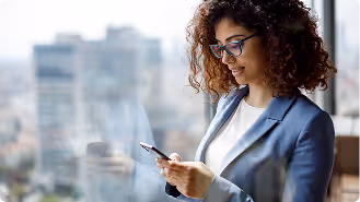Smiling woman with curly hair and glasses using a smartphone near a window with city view.