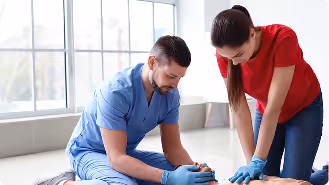 Man in blue scrubs and woman in red shirt performing CPR on a person lying on the floor near a large window.