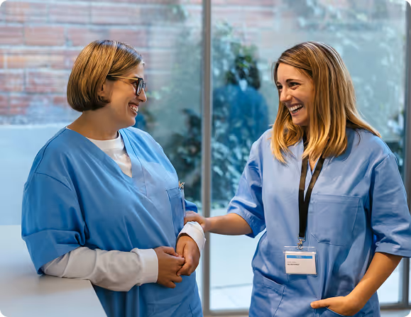 Two female healthcare workers in blue scrubs smiling and conversing in a bright room.