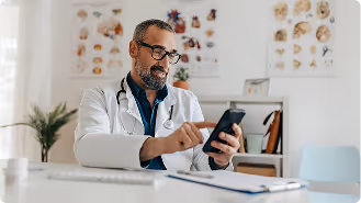 Male doctor in white coat using a smartphone in a medical office with anatomy charts on the wall.