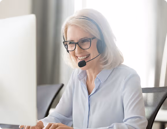 Smiling woman with headset and glasses working on a computer in an office.