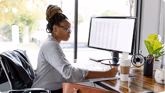 Woman with glasses working on a computer at a wooden desk with a coffee cup and plant.