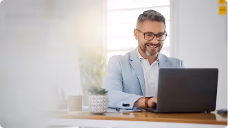 Smiling man in glasses and blazer working on a laptop at a desk with a plant and coffee cup.