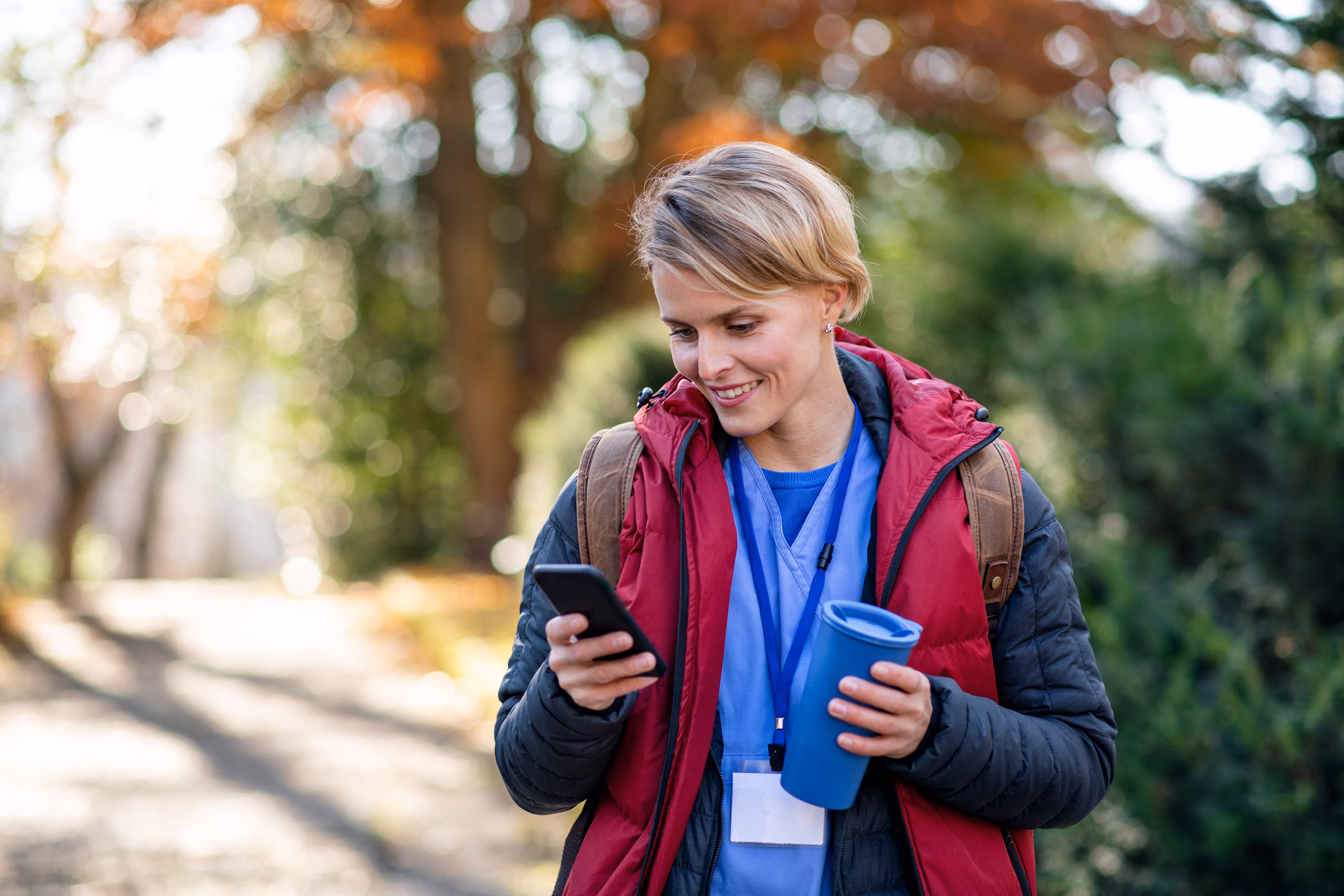 Smiling woman in medical scrubs and red vest holding a blue cup and phone outdoors in autumn.