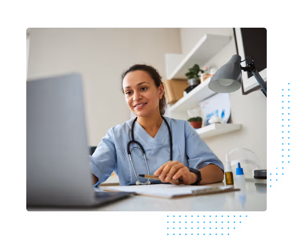 Female healthcare professional in scrubs using laptop and holding a pen at a desk with medical supplies.