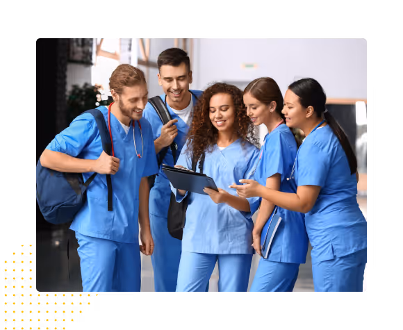 A group of five medical students in blue scrubs smiling and discussing notes on a clipboard indoors.