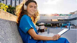 Smiling nurse in blue scrubs sitting outdoors with headphones, coffee cup, and tablet in hand.