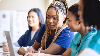 Three diverse women, including one in medical scrubs, working together using a laptop.