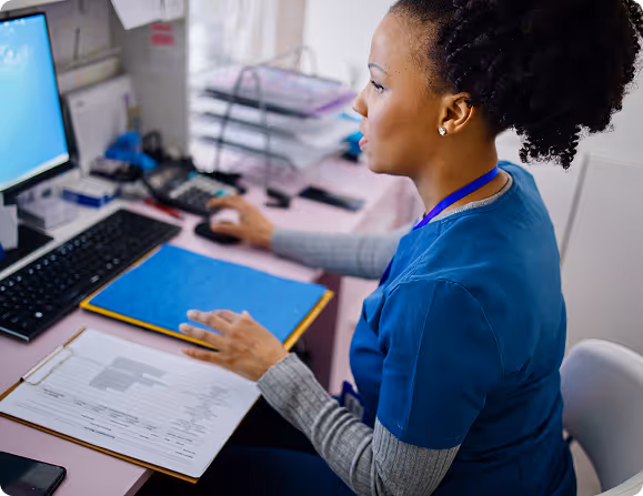 Medical professional in scrubs working at a desk with computer, documents, and a clipboard.