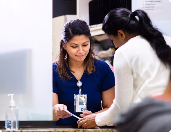 Healthcare worker in scrubs assisting a patient at a clinic reception desk.