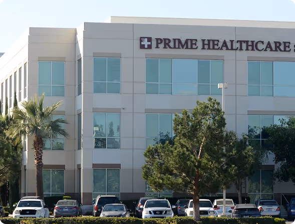 Office building with Prime Healthcare sign, palm tree, pine tree, and parked cars in front.