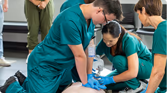 Medical trainees in green scrubs practicing CPR on a manikin during a training session.