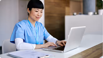Healthcare worker in blue scrubs typing on a laptop at a desk with documents nearby.