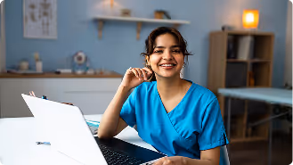 Smiling woman in blue medical scrubs sitting at a desk with an open laptop in a clinic room.