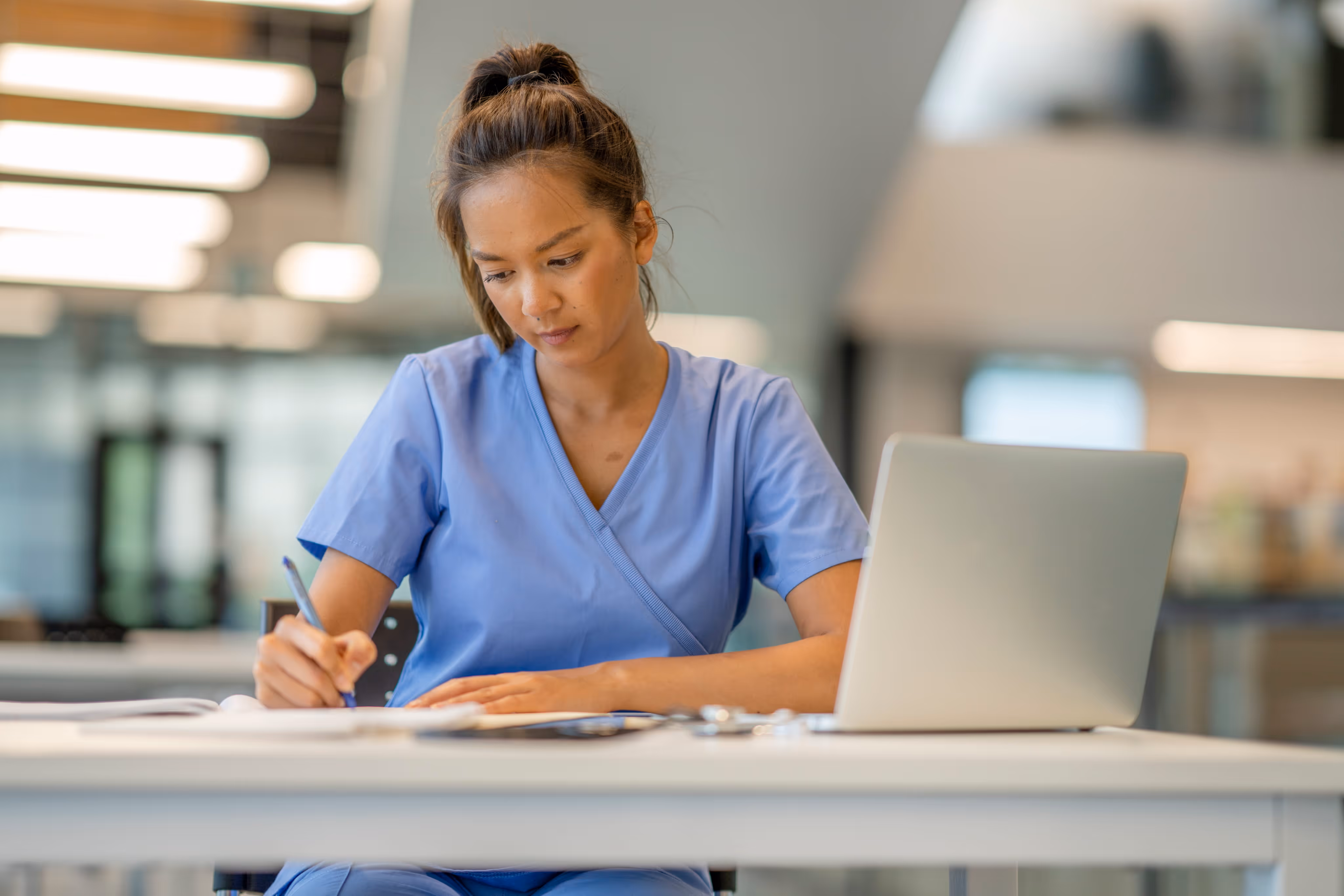 Nurse in blue scrubs writing notes at a desk with a laptop in a modern office setting.