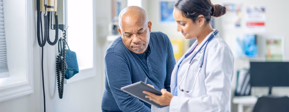 Female doctor with stethoscope consulting middle-aged male patient in medical office with clipboard.