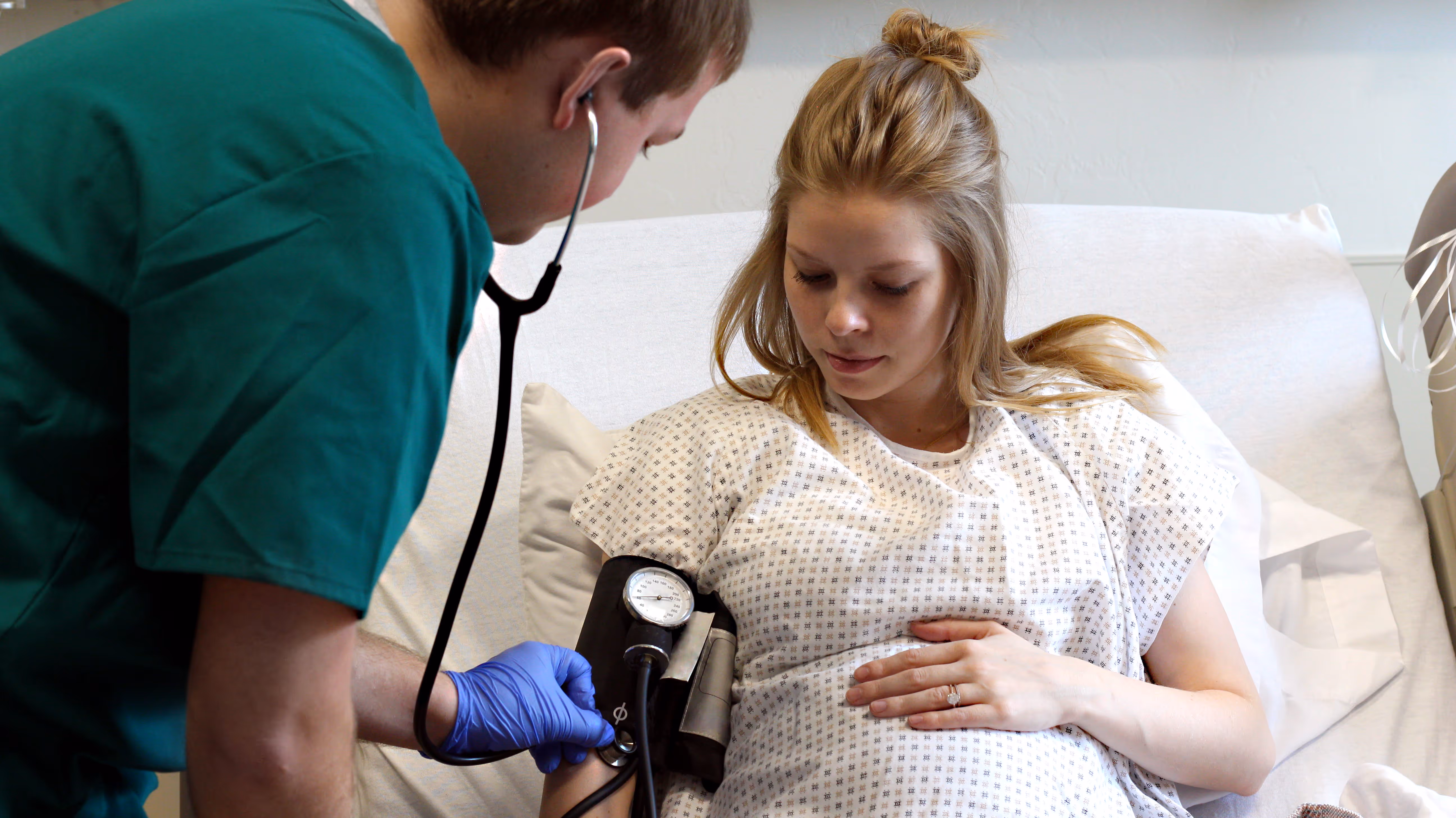 Nurse in green scrubs measuring blood pressure of pregnant woman lying in hospital bed.