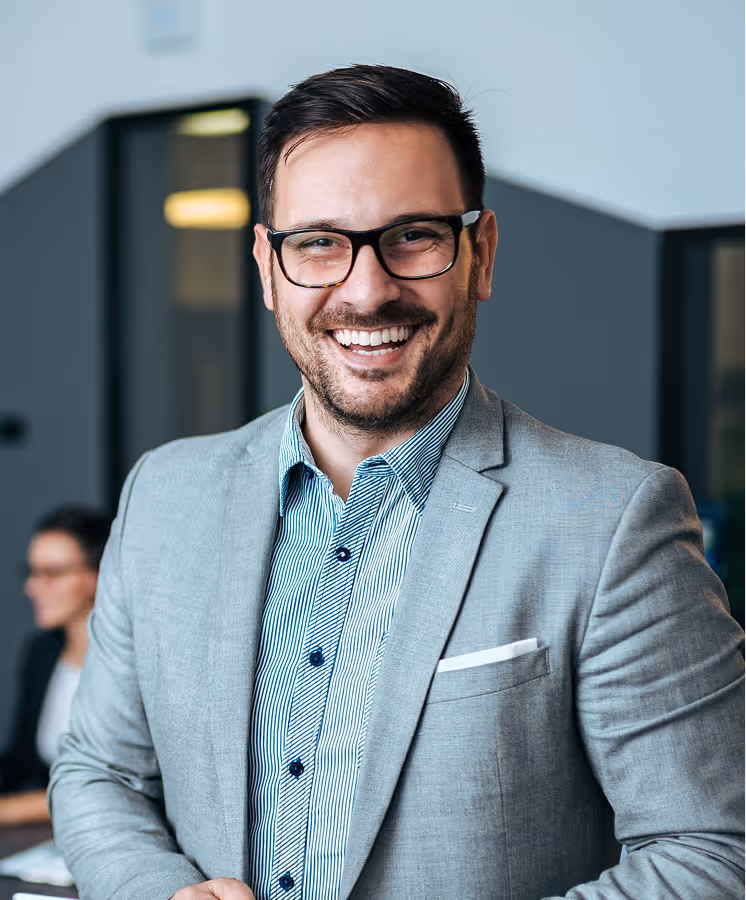 Smiling man wearing glasses and gray suit jacket in a modern office setting.
