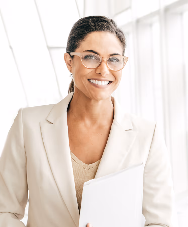 Smiling woman with glasses in beige blazer holding a white folder in a bright office.