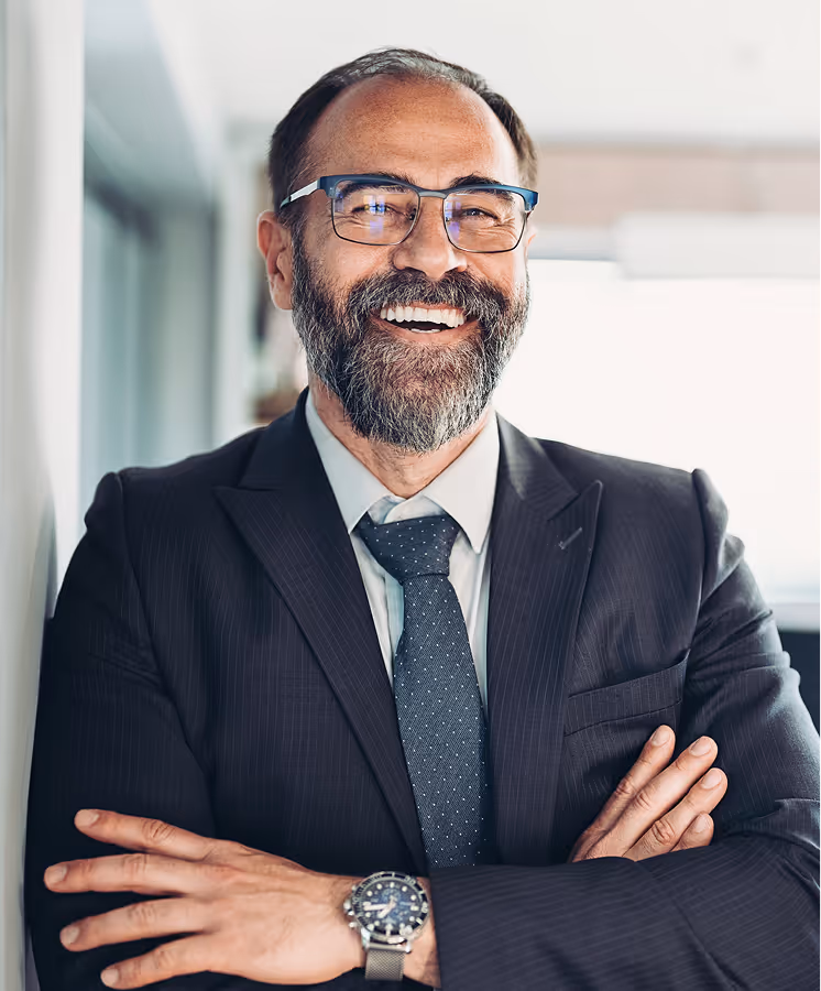 Smiling middle-aged man with glasses, beard, and suit, standing with arms crossed indoors.