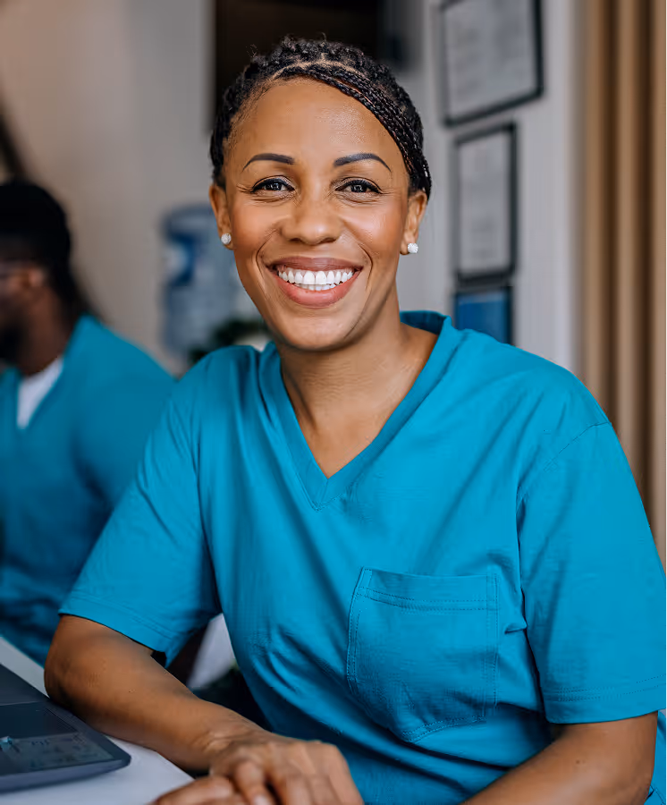Smiling nurse in teal scrubs sitting at a desk with a laptop in a clinical setting.