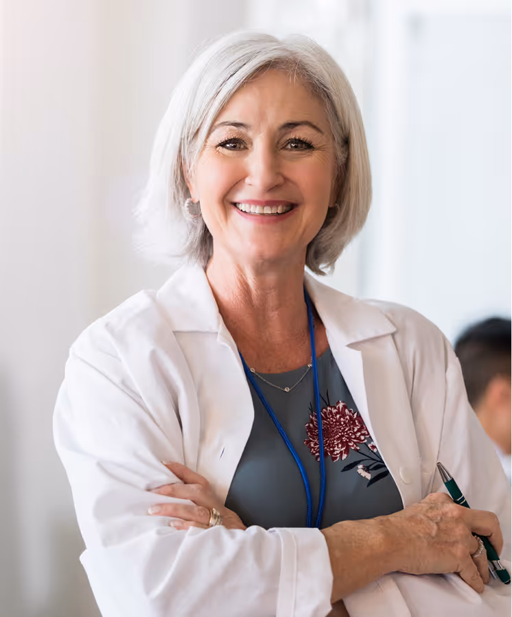 Smiling female doctor with gray hair in white coat holding pen and arms crossed.