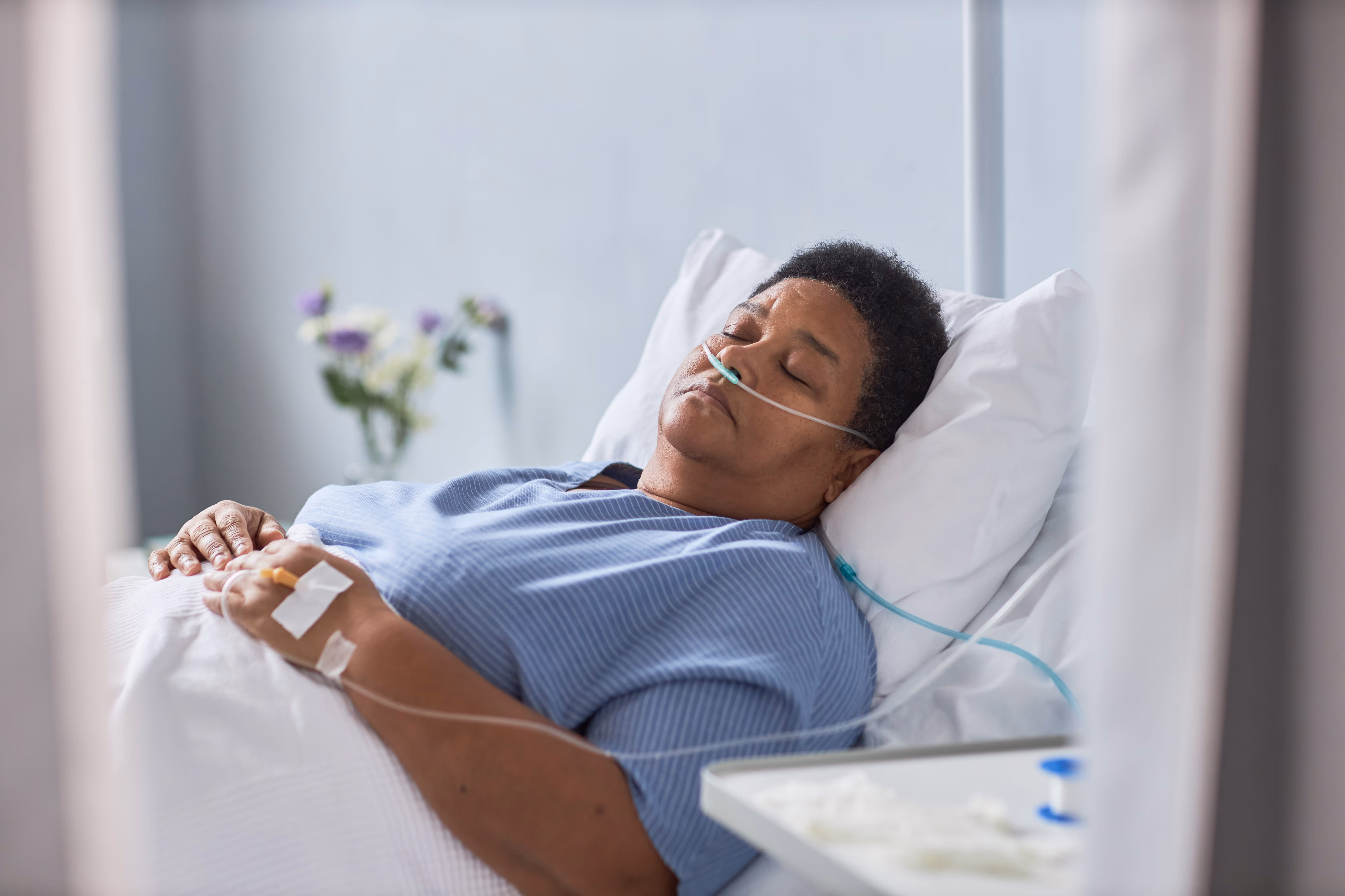 Patient lying in hospital bed with nasal oxygen tube and IV line in hand, eyes closed resting.