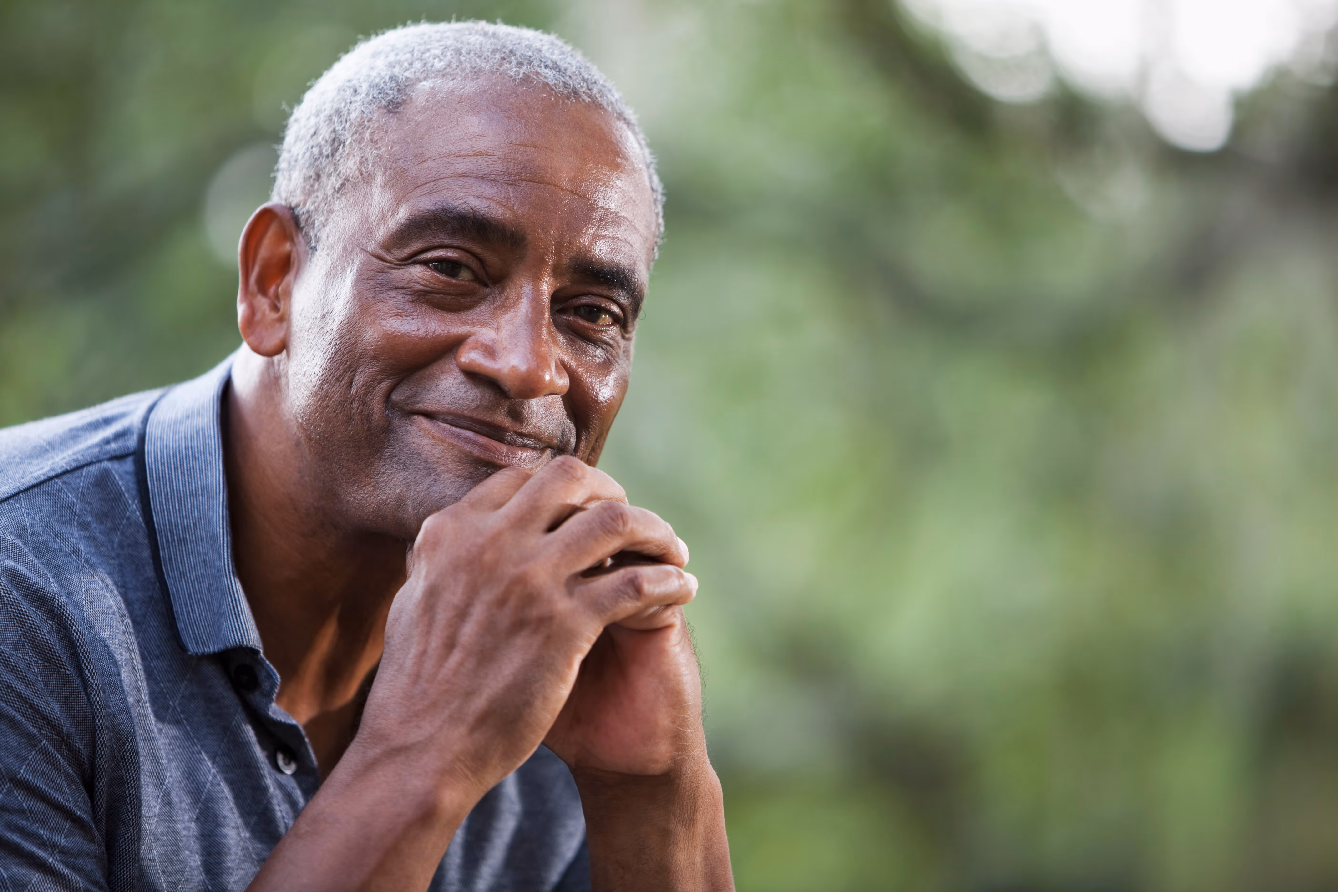Smiling older man with gray hair resting his chin on his clasped hands outdoors with a blurred green background.