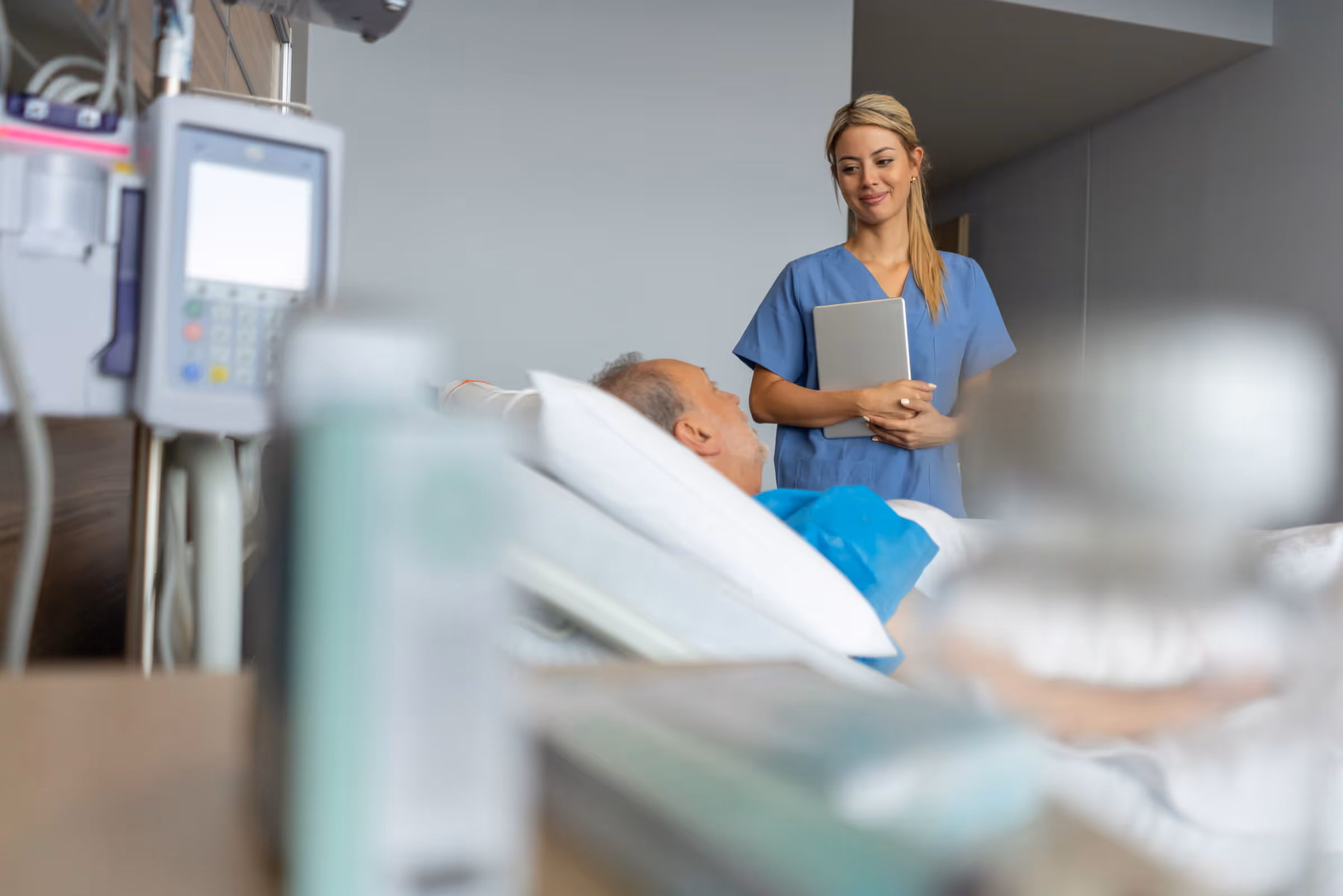 Nurse in blue scrubs holding tablet talking to male patient lying in hospital bed.