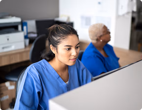 Two medical professionals in blue scrubs working at a desk with a computer in an office.
