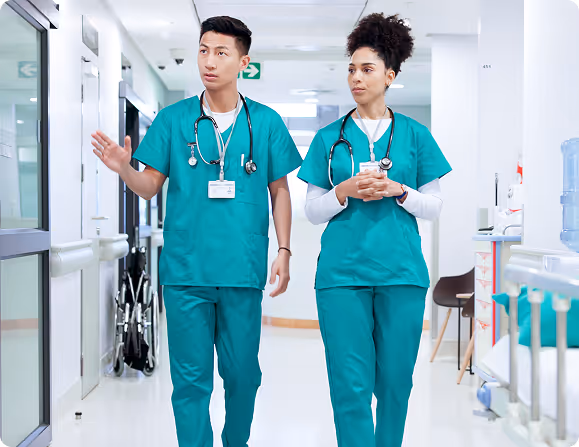 Two healthcare professionals in green scrubs and stethoscopes walk and talk in a hospital corridor.