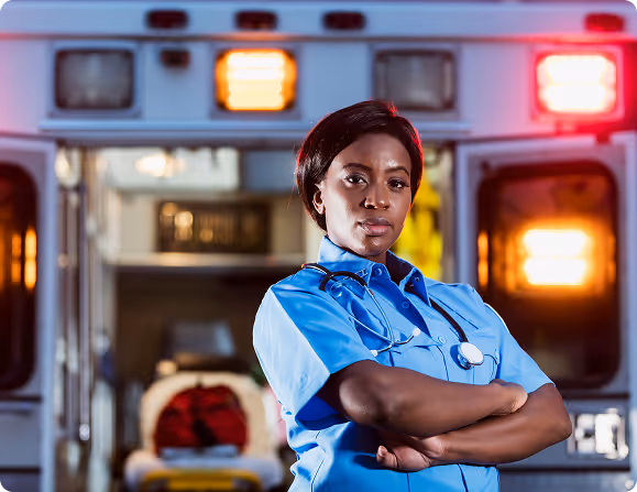 Confident female paramedic in blue uniform with stethoscope stands in front of open ambulance lights on.