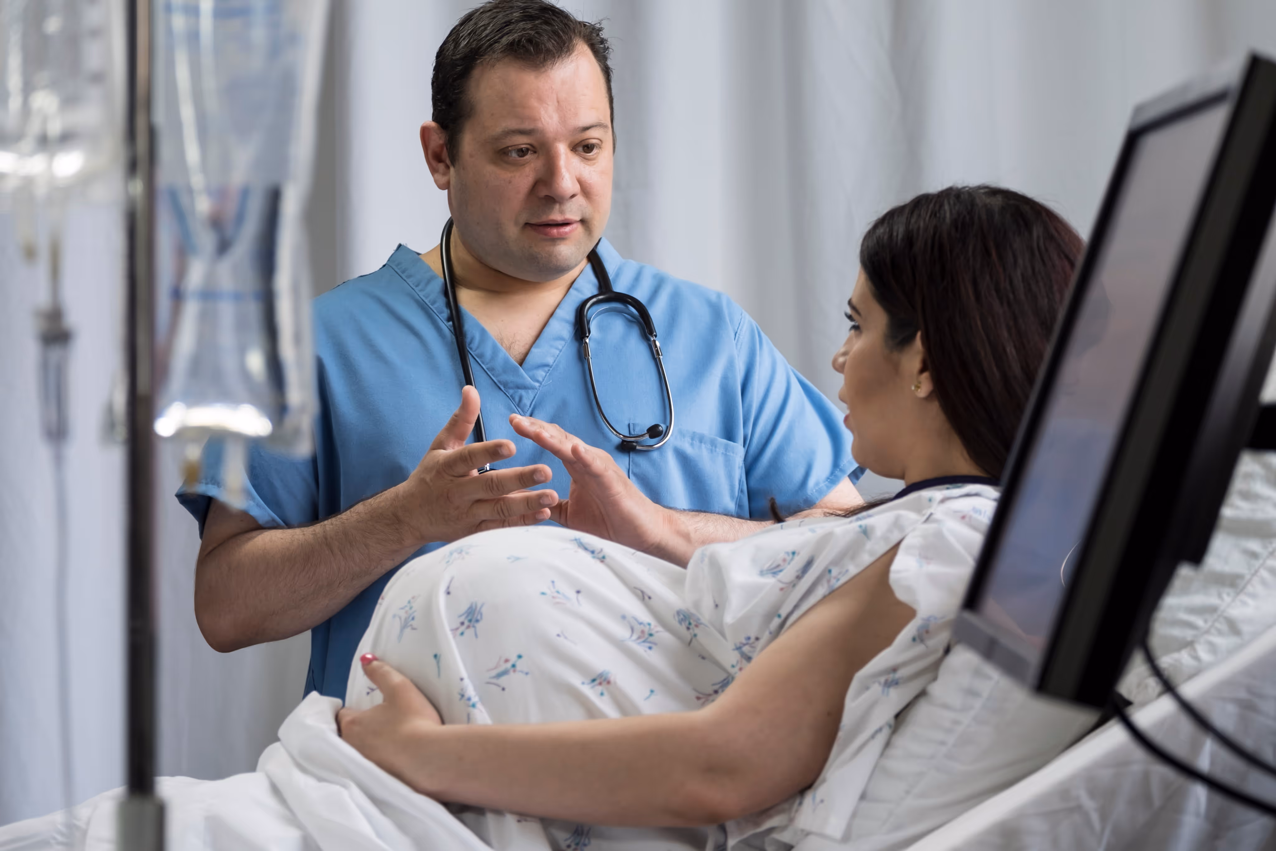 Pregnant woman in hospital gown talking with a doctor in scrubs beside her bed.