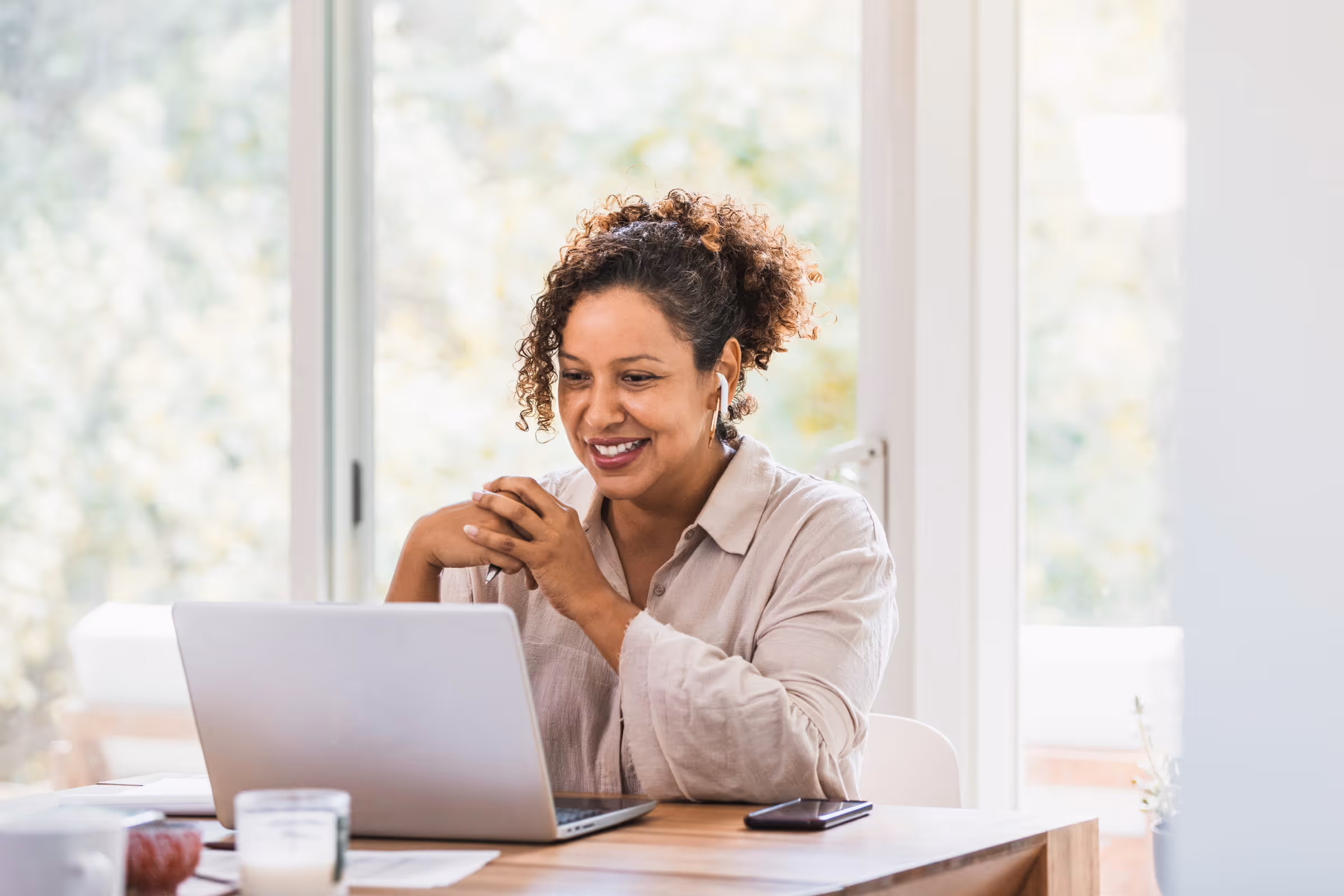 Smiling woman with curly hair using a laptop and wireless earbuds at a wooden table near a window.