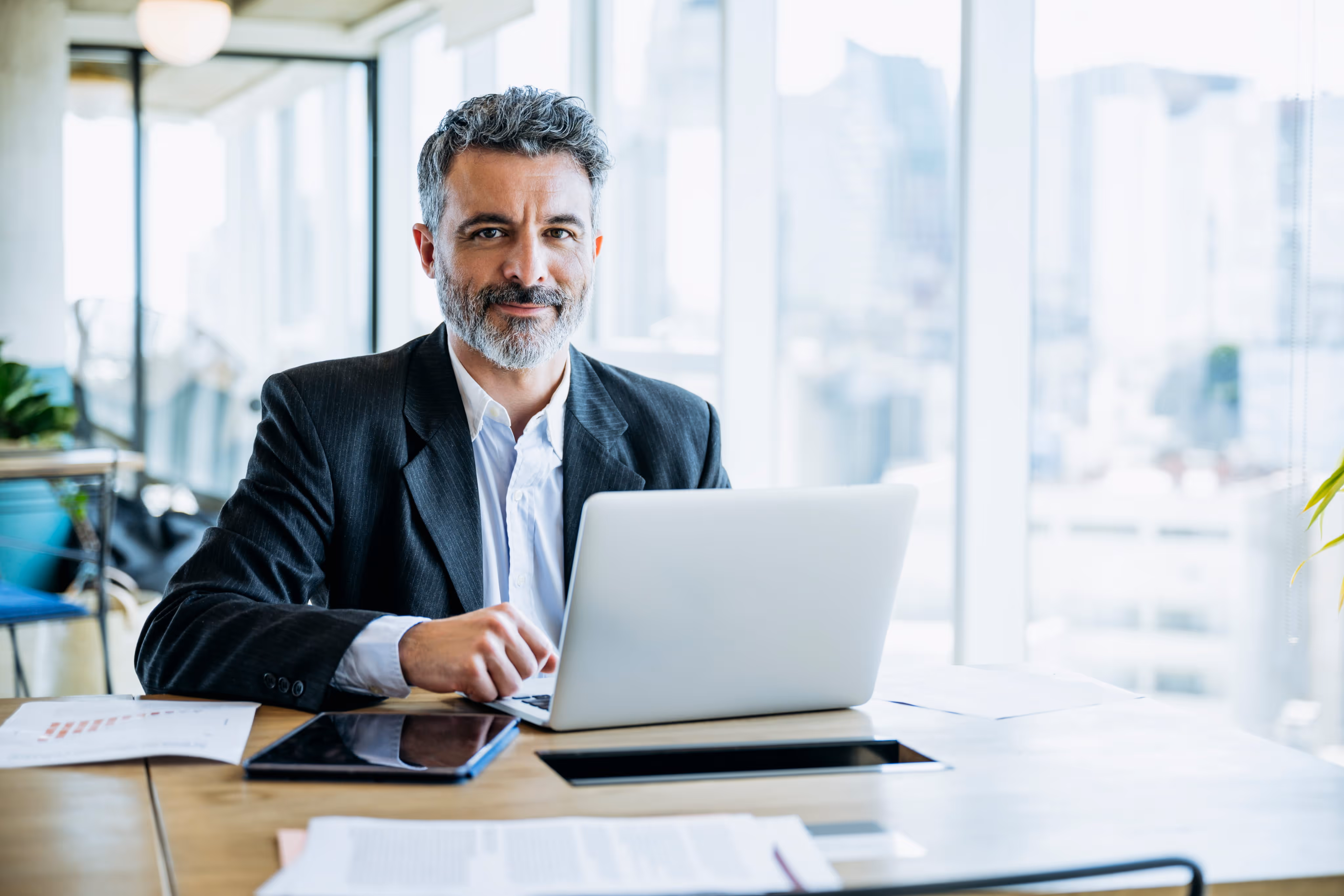 Middle-aged man in business attire working on a laptop in a modern office with city view windows.