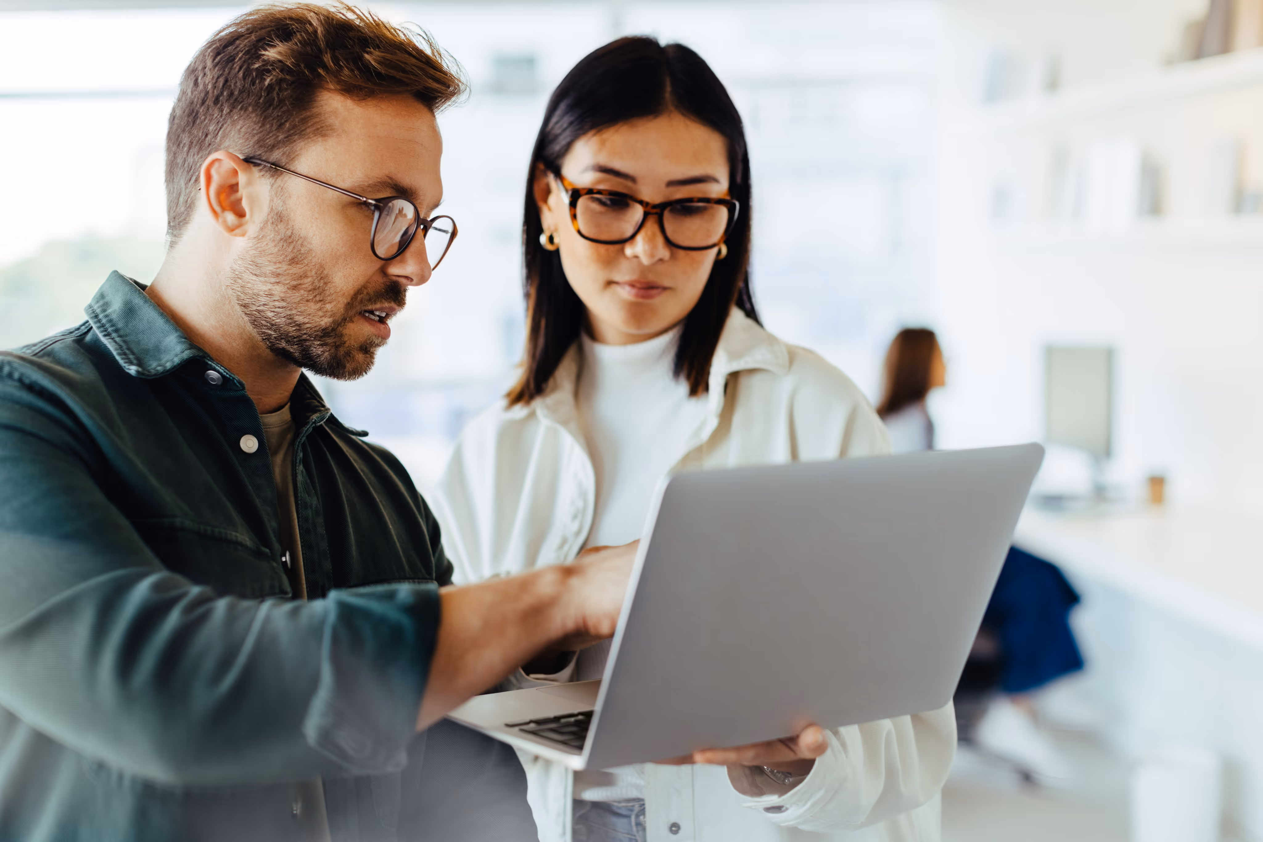 Man and woman wearing glasses looking at a laptop screen in a bright office.