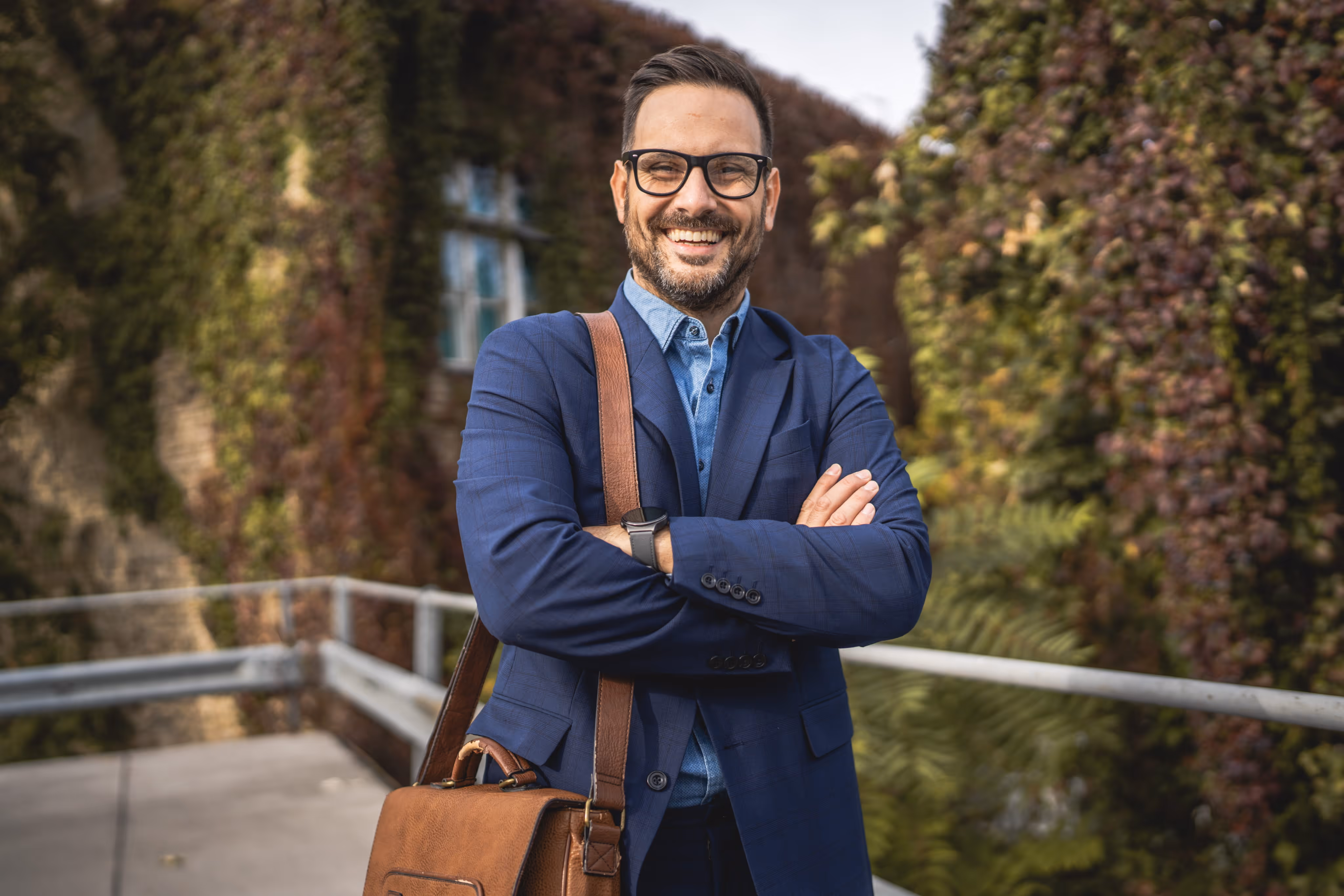 Smiling man with glasses in a blue suit carrying a brown leather bag, standing outside near ivy-covered wall.
